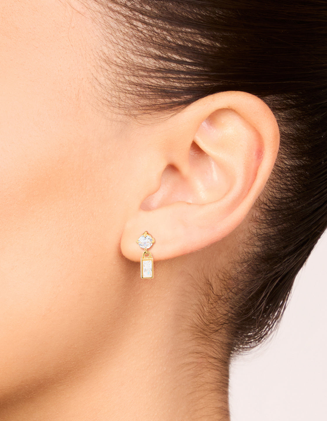 Featuring waterproof drop earrings with stainless steel construction, round and baguette stone cut cubic zirconia in gold plated finish on ear view
