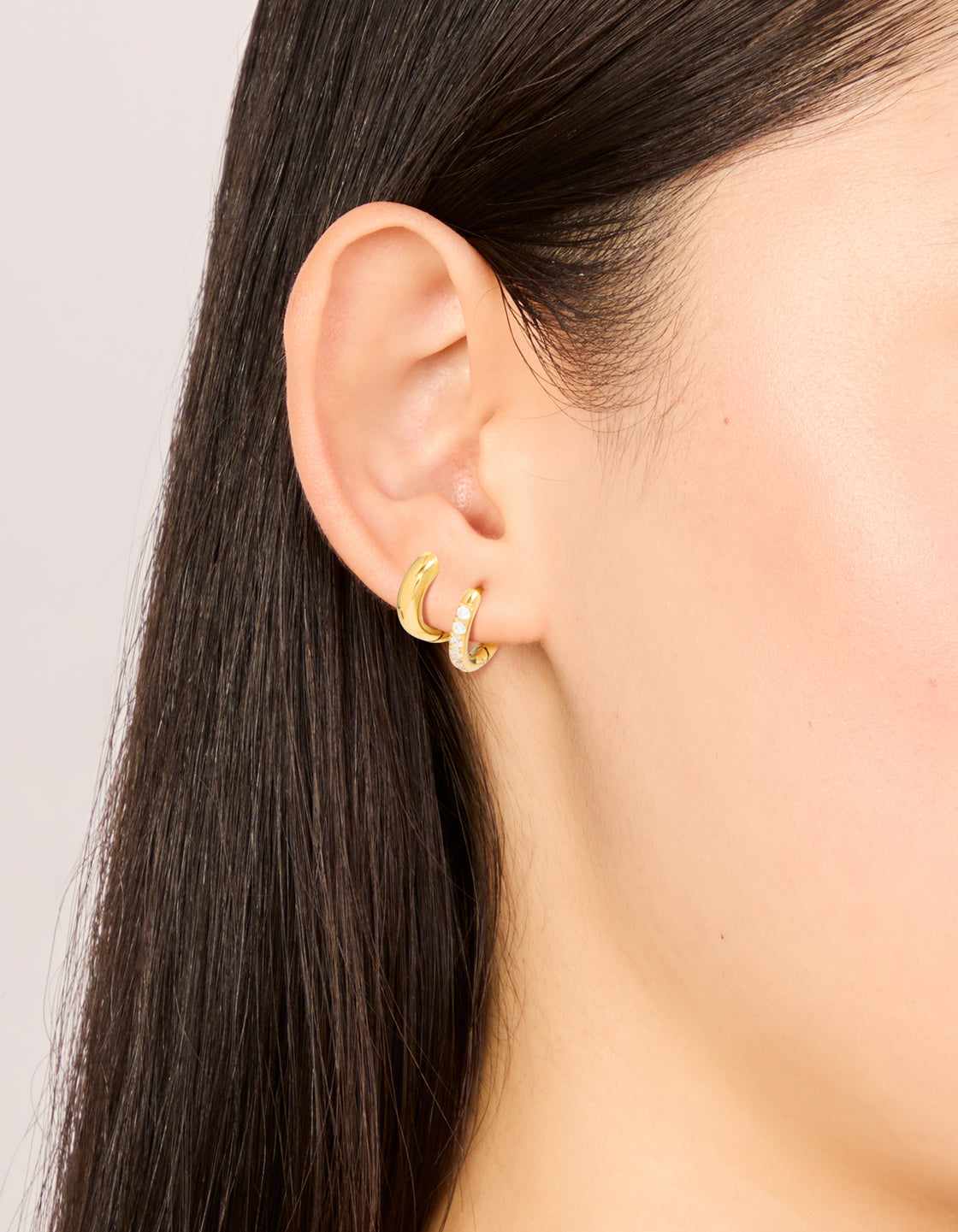 Showcasing waterproof gold plated stainless steel cubic zirconia pavé and plain huggie earrings in a 2-pack on ear view